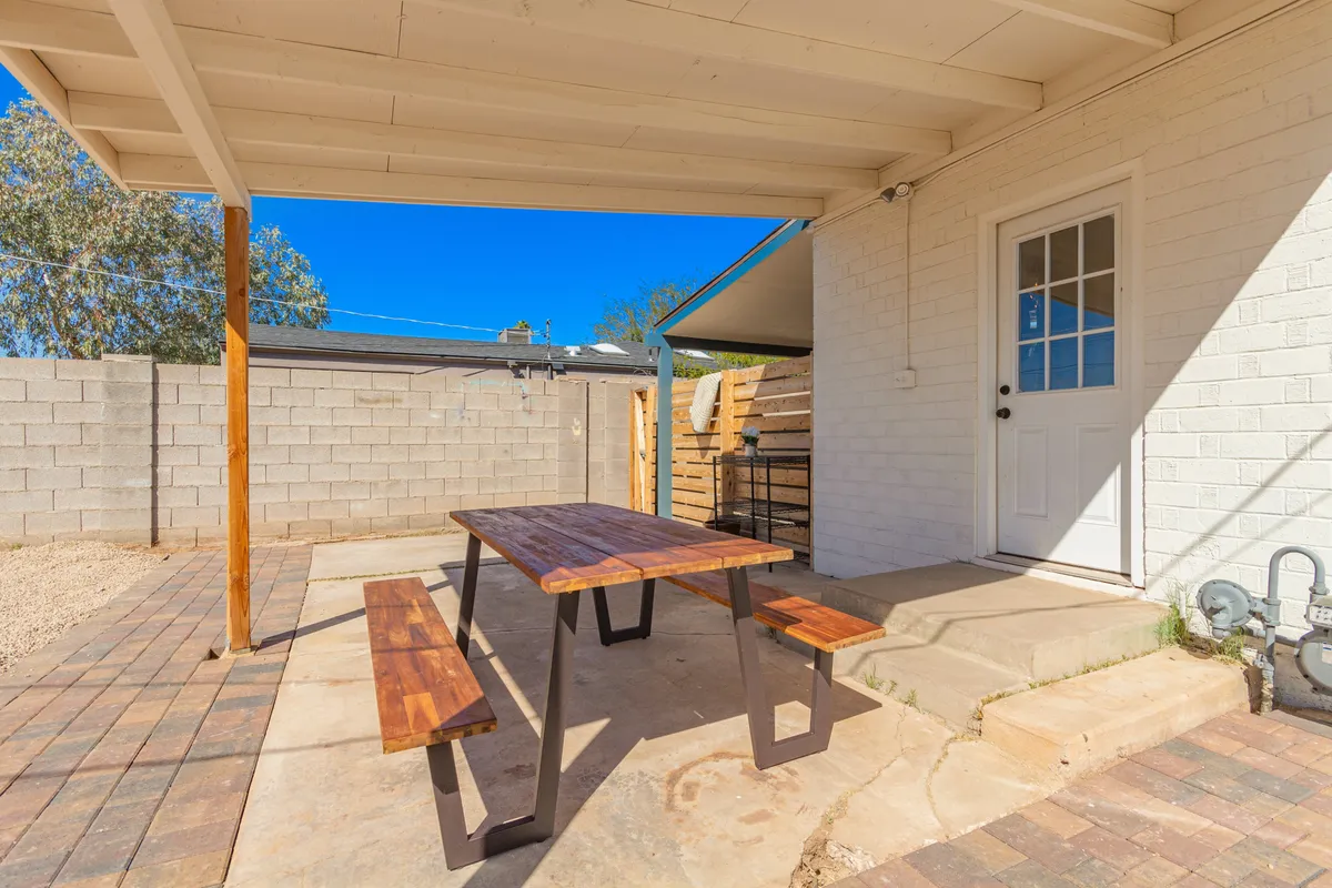A cozy patio space with a wooden table, benches, and a brick wall, ready for outdoor dining or gatherings. The scene feels casual and inviting, suggesting summer barbecues or evening meals under the stars. The space is enclosed for security and privacy, suitable for pets and families.
