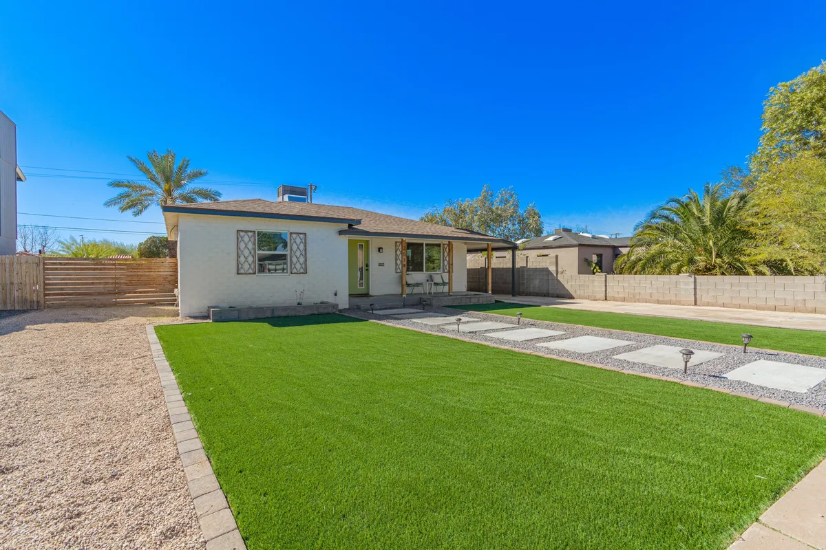 A broad view of a charming house facade with a lush green lawn, paved pathway, and tall palm trees framing the scene. The property is set back from the street, emphasizing the neighborhood's mature landscaping and relaxed, welcoming vibe. The sunlight accentuates the classic architectural lines and the inviting outdoor spaces ideal for strolls or socializing.