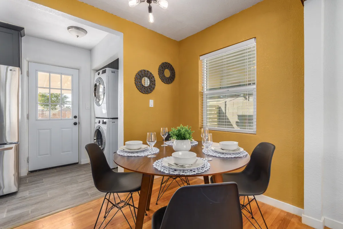 A cozy dining area centered around a round table elegantly set with plates and glasses, with a prominent yellow accent wall adding warmth. The background reveals a laundry area, hinting at the home's practical layout. The scene exudes an inviting atmosphere perfect for relaxed family meals or intimate gatherings.