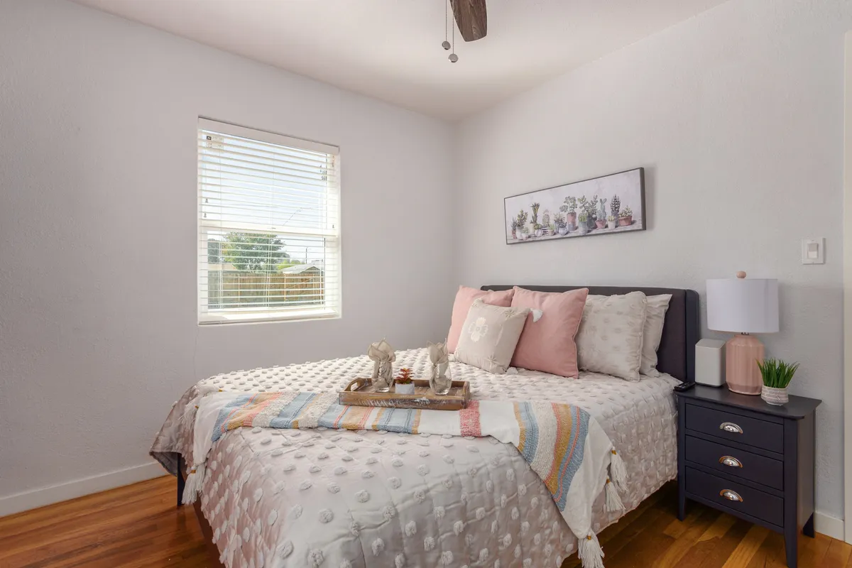 A calm, serene bedroom with a comfortable bed adorned with decorative pillows and soft blankets. A nightstand with a lamp adds warmth, while wall art above the bed and a window with blinds show attention to detail and provide gentle natural light.