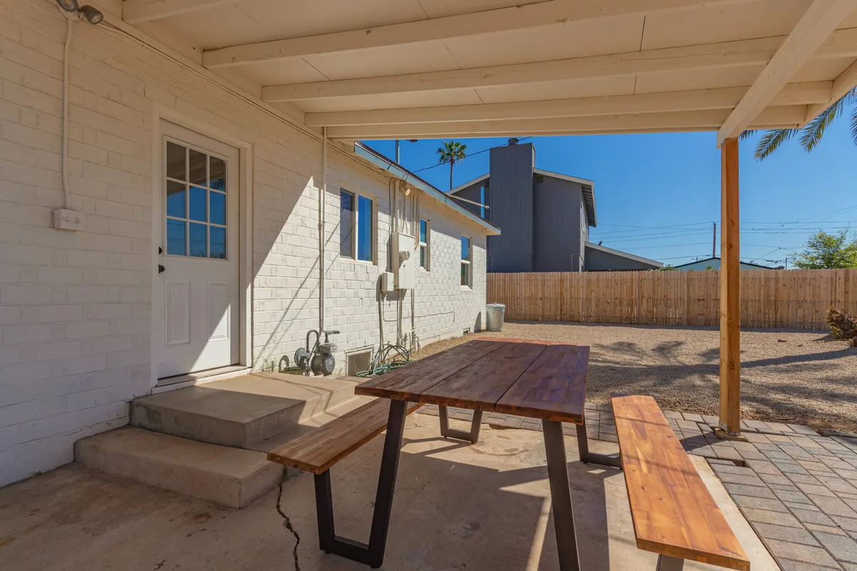A cozy patio area featuring a wooden table set for outdoor dining, shaded space, and a welcoming doorway. The area is designed for casual gatherings or quiet moments, surrounded by natural greenery, offering a perfect spot to recharge at home.