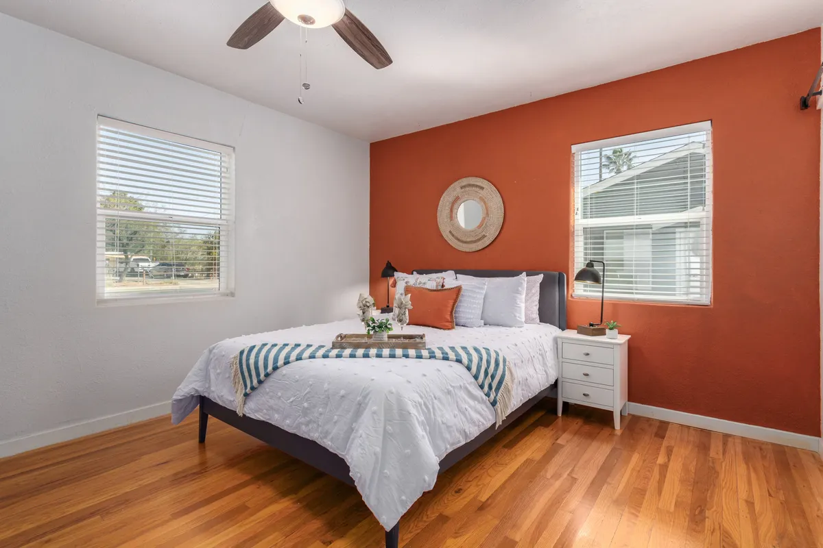 A tranquil bedroom featuring a neatly made bed with soft decorative pillows, set against a soothing orange accent wall. A decorative mirror and a nightstand with a warm lamp create a cozy ambiance, with natural light filling the space.
