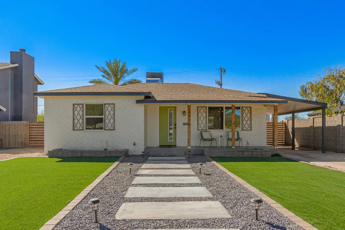 A front-facing exterior shot featuring a sunlit green lawn, a welcoming front door, and large windows. The pathway leads directly to the entrance, framed by mature greenery and trees, conveying a sense of arrival and community. The porch and windows hint at leisurely mornings and evenings spent outdoors.