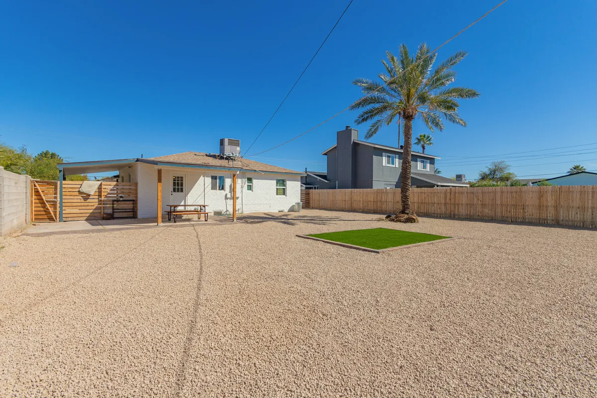 This exterior shot captures a relaxed backyard scene featuring a palm tree, a gravel yard interspersed with a green grass patch, and children playing in the background. The back of the house acts as a welcoming backdrop, while the open space suggests a place for family gatherings and playful afternoons.