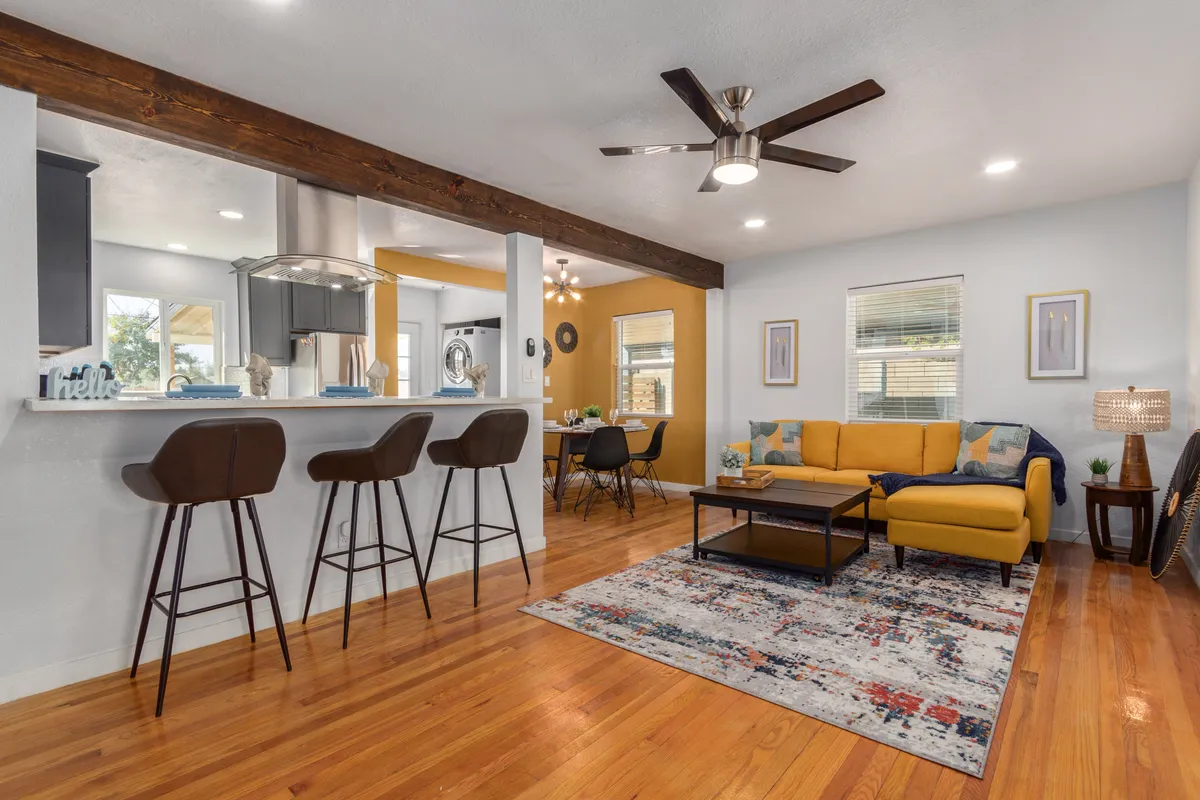 This image captures a welcoming living room with a sunny yellow sofa, visible wooden beam, bar stools at a counter, and a textured area rug. The space is accented with modern fixtures and is filled with natural light, highlighting both contemporary and vintage-inspired elements.