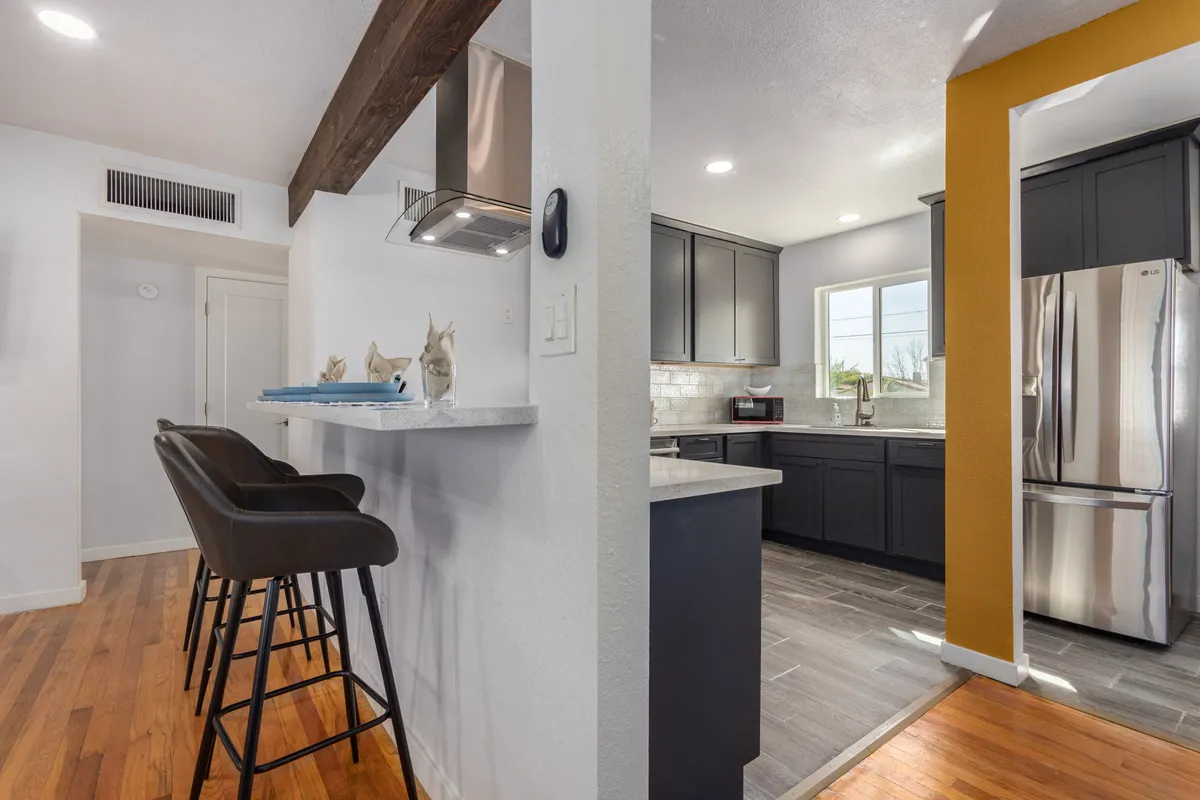 A welcoming kitchen scene featuring stylish bar stools lined up at the kitchen island, stainless steel appliances gleaming under the light, and a cheerful yellow accent wall in the background. The space is well-lit and designed for both cooking and socializing, with ample countertop space and seating that encourages interaction.