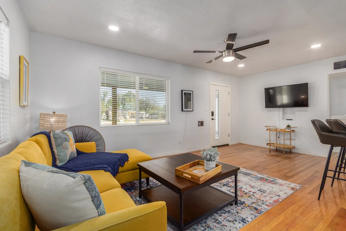 A sunlit living room with a yellow couch, central coffee table, wall-mounted TV, and a window streaming in natural light. The space is cheerful, with modern and classic touches blended seamlessly, ideal for unwinding or hosting friends.