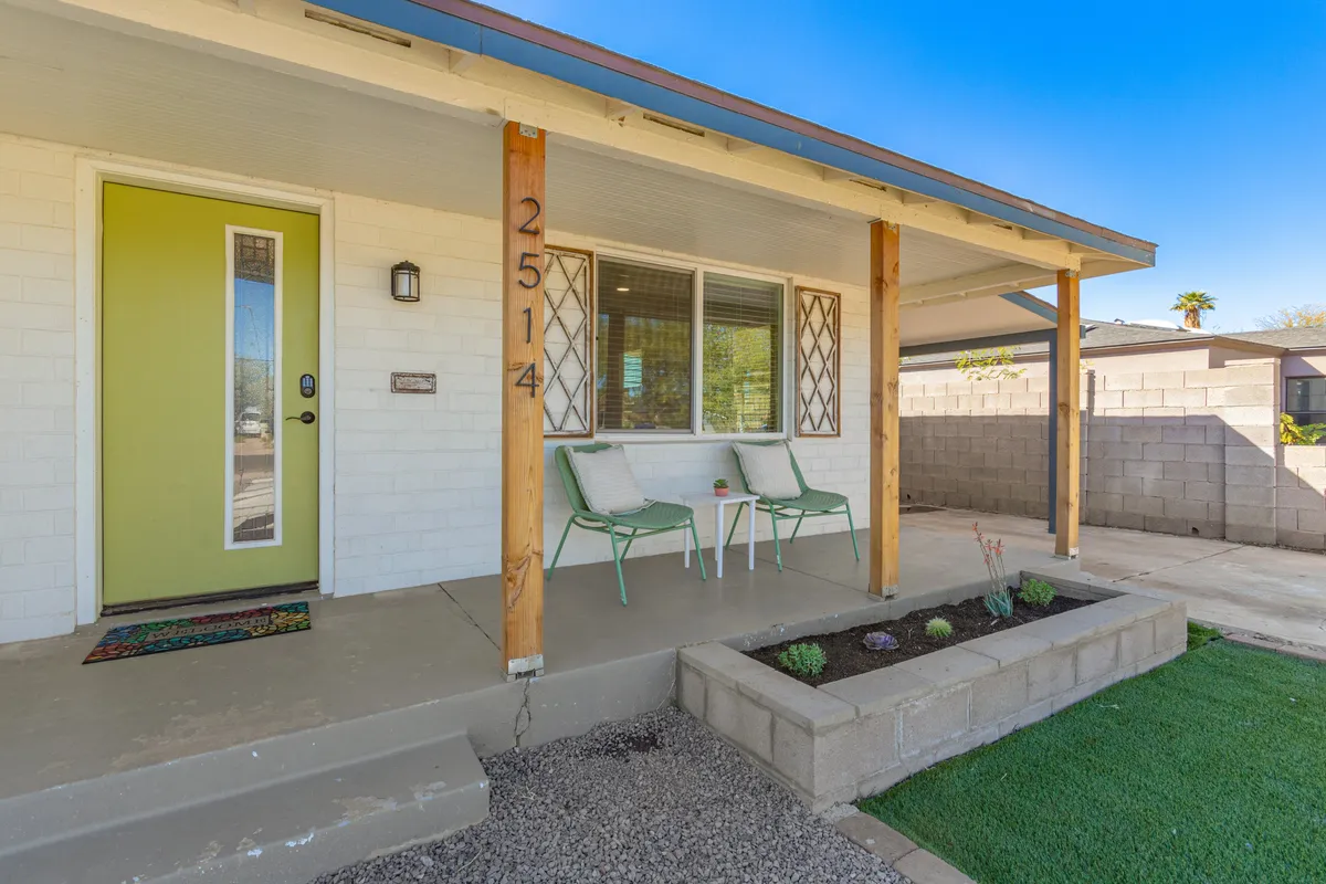 A stylish front porch with outdoor seating, a distinctive green front door, and a decorative house number. The scene feels intimate and approachable, suggesting moments spent relaxing, sipping coffee, or socializing while watching the neighborhood activity.